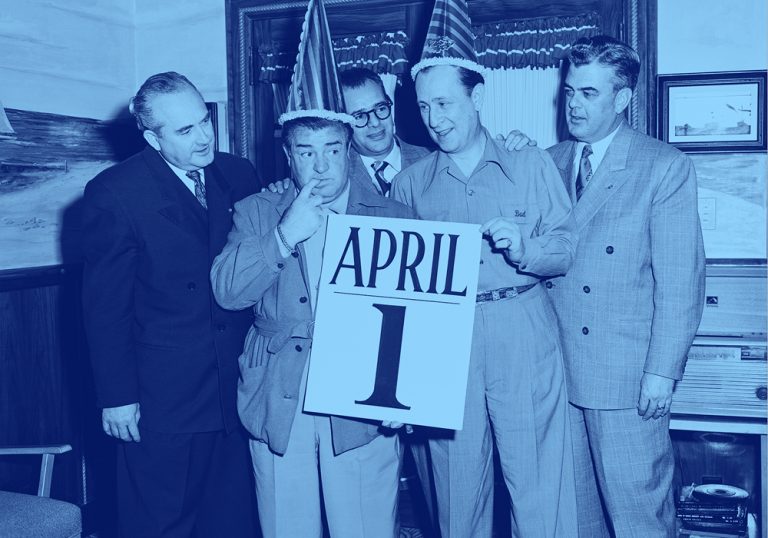 two men in party hats hold a giant calendar page reading "April 1st"