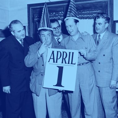 two men in party hats hold a giant calendar page reading "April 1st"