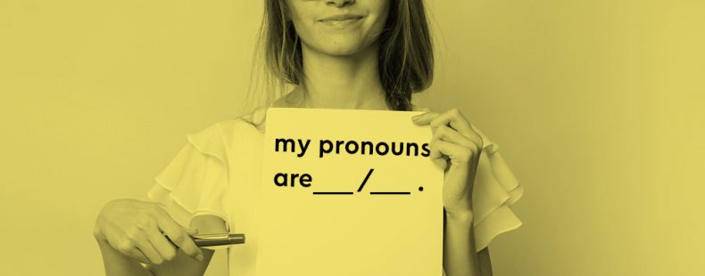 Young woman holding up and pointing to a paper that says, "my pronouns are ___/___", yellow filter.