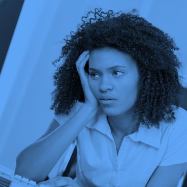 image of woman with her head in her hand sitting at a desk at work, blue filter.